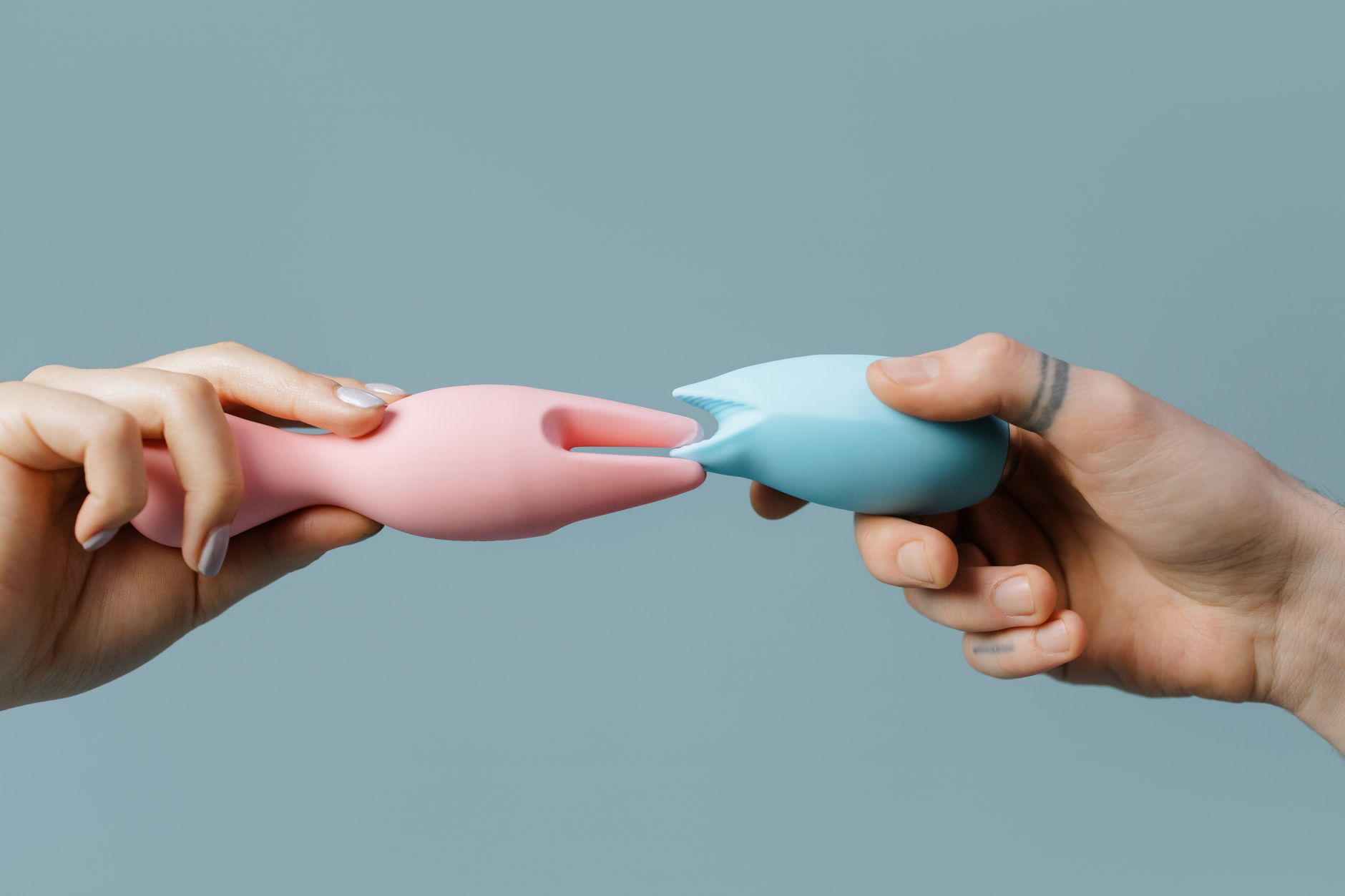 Zwei Hände halten rosa und blaue Silikonvibratore gegen einen Pastellhintergrund