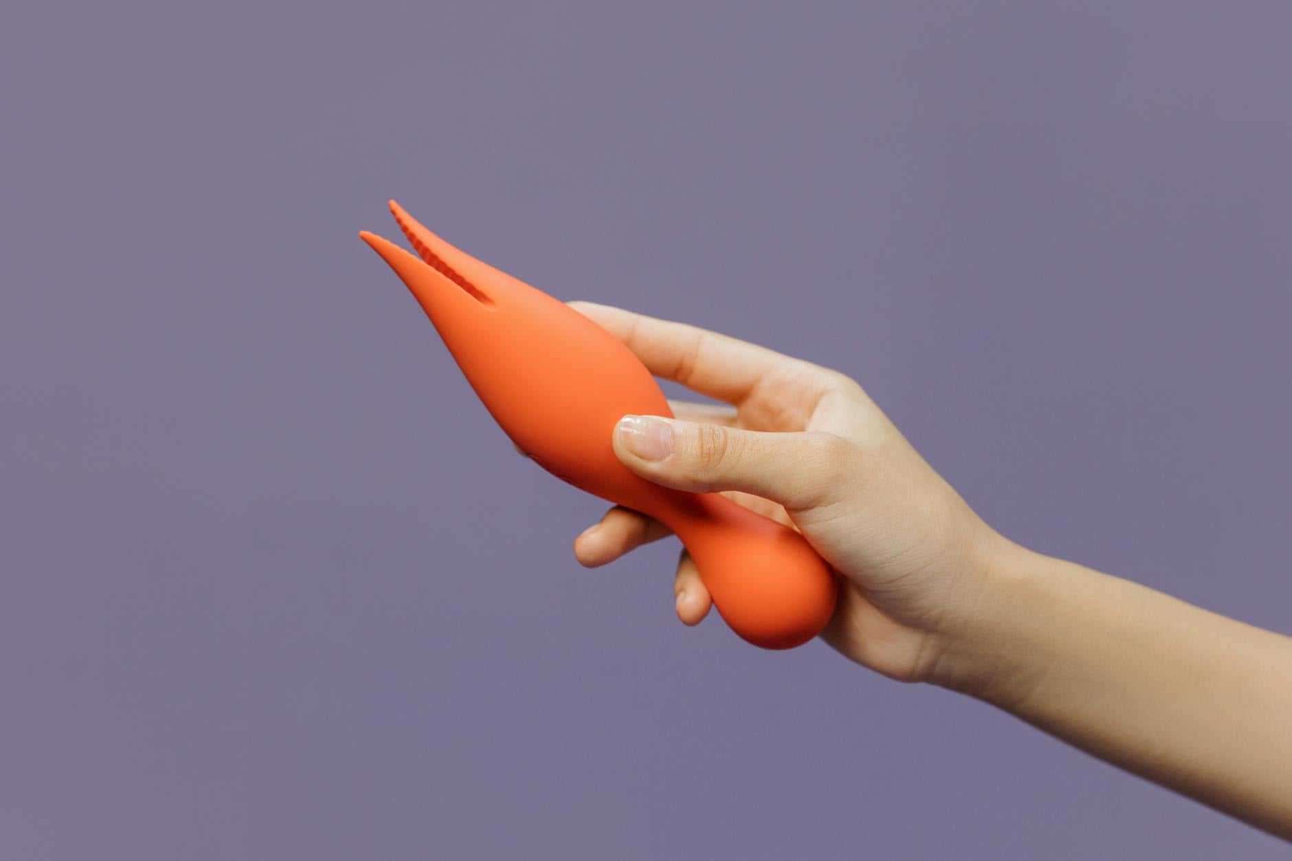Eine Hand hält einen orangefarbenen Vibrator vor minimalistischem lila Hintergrund