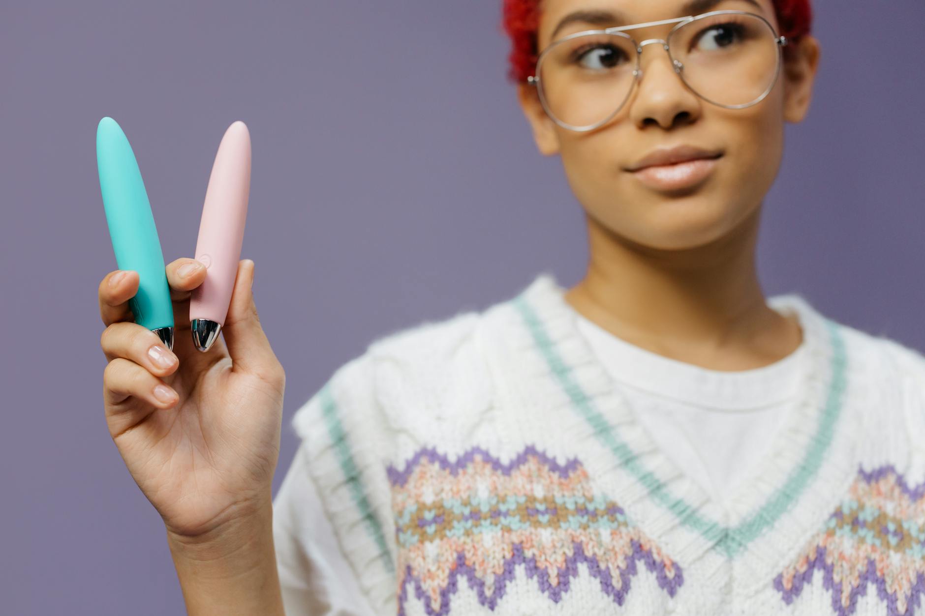 Junge Frau mit Brille hält zwei farbenfrohe Vibratoren vor violettem Hintergrund